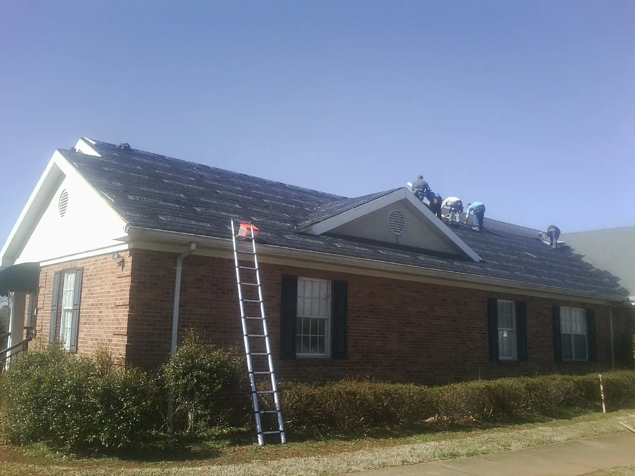 Crew tearing shingles off a brick ranch roof with a ladder set — Metal Roof Repair work by our crew in Mumford