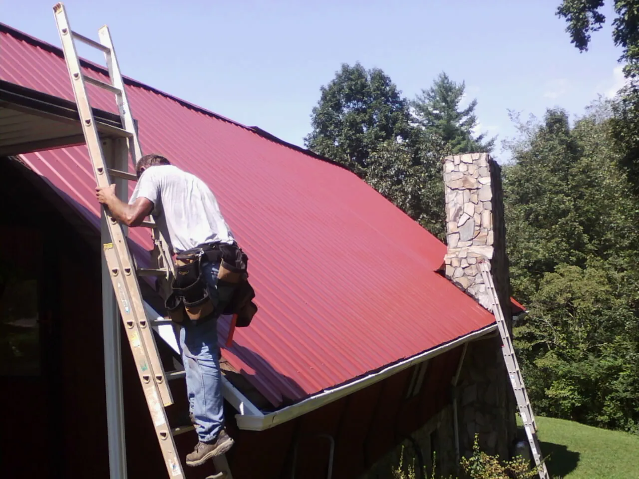 Roofer on a ladder beside a red metal roof and stone chimney — Soffit & Fascia Repair by Roofing Brothers in Mumford